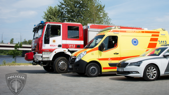 Pie upes novietoti stāvēšanai cits citam blakus operatīvie transportlīdzekļi, priekšplānā policijas mašīna, pa vidu neatliekamās medicīniskās palīdzības dienesta mašīna, tālāk valsts ugunsdzēsības un glābšanas dienesta mašīna