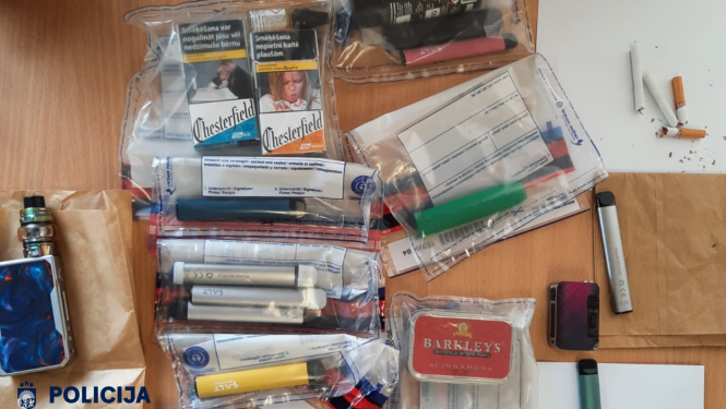 Uz galda dažādas elektroniskās smēķēšanas ierīces, cigarešu paciņas, uzpildes šķidruma tvertnes un cigaretes