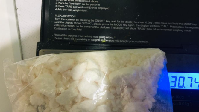 Caurspīdīgā plastmasas maisiņā balta viela uzlikta uz elektroniskajiem svariem, kas rāda 30,74 gramus