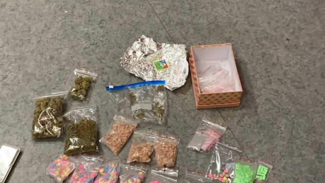 Uz grīdas nolikti vairāki caurspīdīgi plastmasas maisiņi ar dažādu krāsu tabletēm - narkotikām, marihuānu, folijā ietītas markas, pelēks mobilais tālrunis