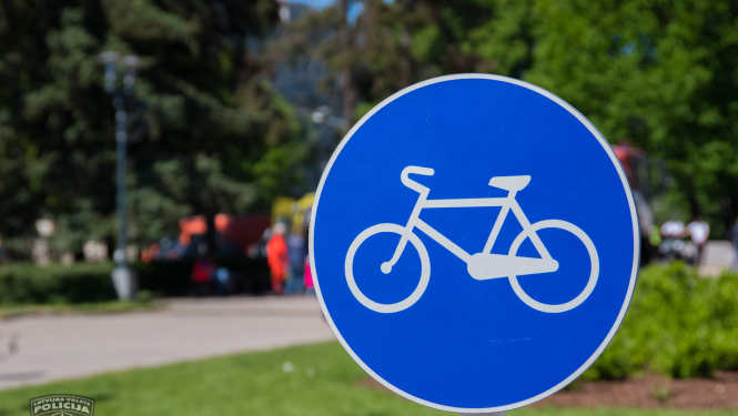 Ceļa zīme, kas norāda riteņbraucēju ceļu, fonā redzams parks