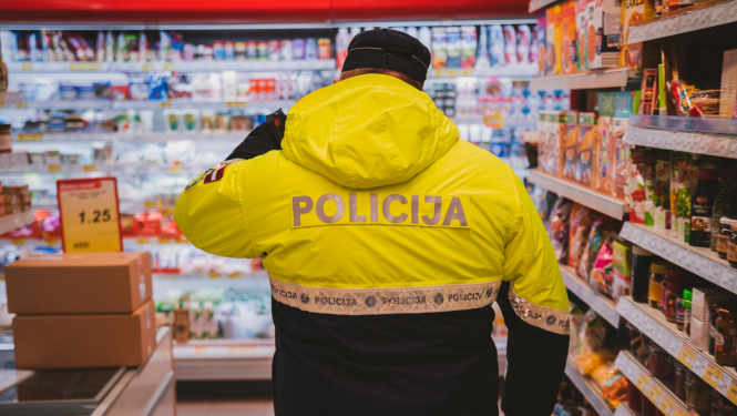 Policists formā no mugurpuses, atrodas veikalā. Vienā malā plaukti, priekšā fonā redzama piena produktu nodaļa