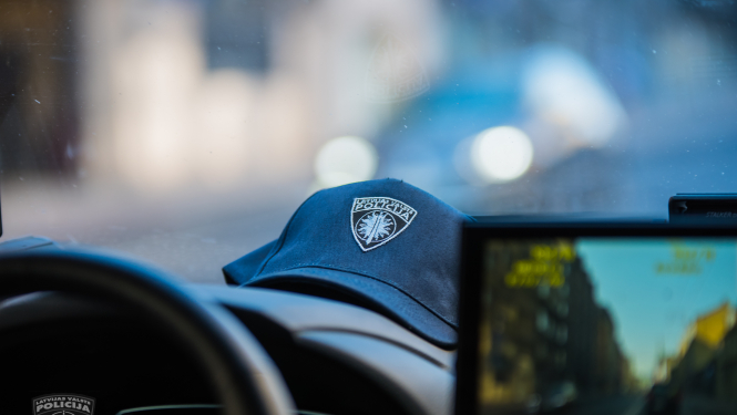 Policijas automašīnas iekšpuse, redzama stūre, daļa no ekrāna, uz priekšējā paneļa uzlikta policista cepure