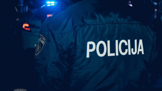 Policists formā, uz muguras uzrakstīts Policija, tālumā fonā policijas mašīna ar ieslēgtām bākugunīm, tumšajā diennakts laikā