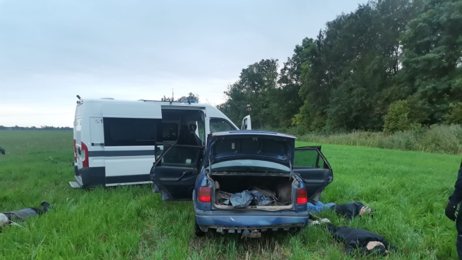 Policijas automašīna piebraukusi priekšā vieglam auto, kam visas durvis atvērtas, arī bagāžas nodalījums. Blakus automašīnai zālē guļ 3 vīrieši ar seju pret zemi