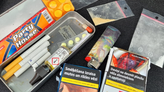 Metāla penālis, kurā atrodas cigaretes, tabletes, žilete, pie penāļa cigarešu paciņas, caurspīdīgi plastikāta maisiņi ar baltas un oranžas krāsas pulverīšiem