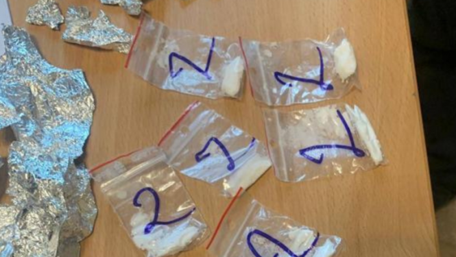 Uz galda vairāki caurspīdīgi plastmasas maisiņi, kuros iekšā balta pulverveida viela, un folijas loksnes