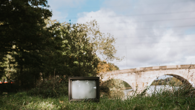 Vecs televizors novietots zālē, fonā redzami krūmi un akmens tilts ar arkām; saulaina diena; vasara