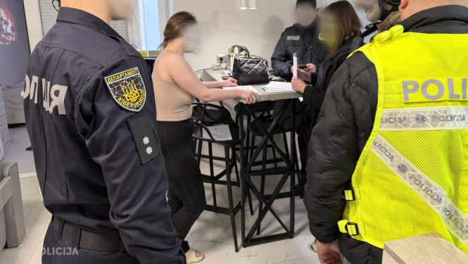 Valsts policijas darbinieki dzīvoklī veic procesuālās darbības, vairāki policisti formas tērpos atrodas pie galda, kur sēž sieviete, fonā redzama moderna virtuves zona