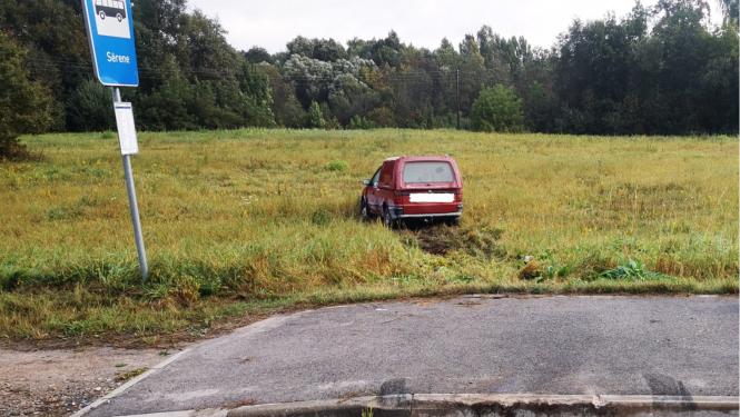 Sarkana automašīna iebraukusi pļavā, priekšplānā autobusa pietura