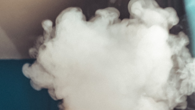 Cilvēka rokā tiek turēta melna elektroniskās smēķēšanas ierīce, seju sedz dūmu mākonis