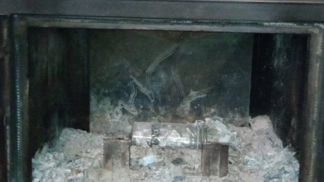 Kamīns, kurā redzami pelni, metāla gabals un arī stikla burka, kurā ielikti folija vīstoklīši