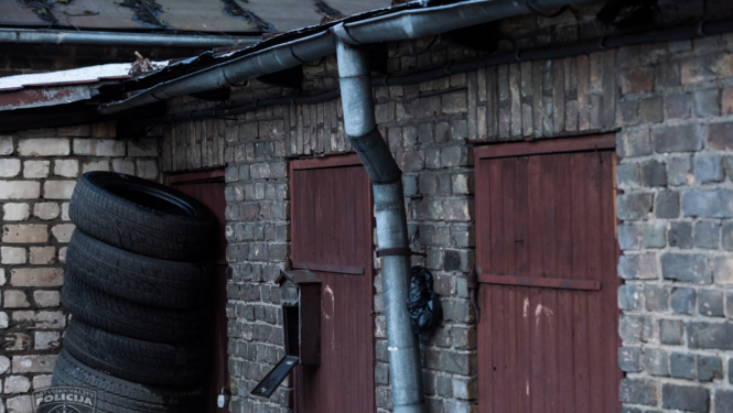 Ķieģeļu ēka, trīs brūnas koka ārdurvis, vienām durvīm priekšā sakrautas automašīnu riepas