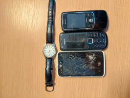 uz gaišas koka imitācijas virsmas novietoti rokas pulkstenis, viedtālrunis un divi podziņtelefoni