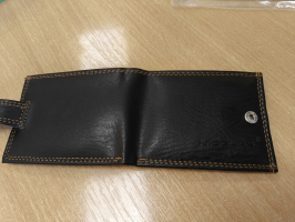 Atvērts melns ādas maks ar brūnām nošuvēm un metāla spiedpogu, novietots uz gaiša koka galda. Labajā pusē ādā iespiests zīmola nosaukums "Nicolas"