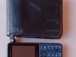 Nobružāts melns ādas maks, kas novietots tieši virs tumši zila "Nokia" podziņtelefona uz gaišas koka virsmas.