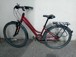 Tumši sarkans (bordo) Kross zīmola pilsētas velosipēds ar pazeminātu rāmi. Velosipēds ir aprīkots ar melnu sēdekli, aizmugurējo bagāžnieku, dubļusargiem, atbalsta kājiņu un zvaniņu uz stūres. Tas novietots ārā uz asfalta, atbalstīts pret gaišu, teksturētu sienu.