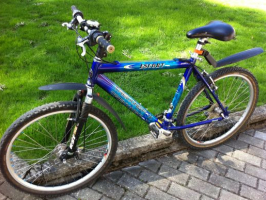 Tumši zils kalnu velosipēds "Escort Trekking Fox" ar priekšējo amortizāciju un melniem dubļusargiem, atbalstīts pret zemu bruģa apmali blakus zaļam zālienam.