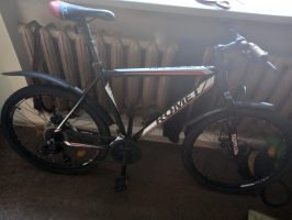 melns 'Romet Rambler' kalnu velosipēds ar baltiem un sarkaniem dizaina elementiem. Aprīkots ar priekšējo amortizatoru, disku bremzēm un melniem plastmasas dubļusargiem