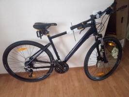 Melns "Scott" kalnu velosipēds ar dzelteniem akcentiem, aprīkots ar priekšējo amortizāciju un disku bremzēm, atbalstīts pret baltu sienu iekštelpās uz koka grīdas