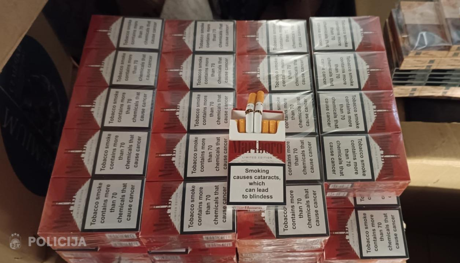 liels daudzums cigarešu paciņu, kas sakrautas kastē vairākās rindās, virspusē novietotas arī dažas atsevišķas cigaretes