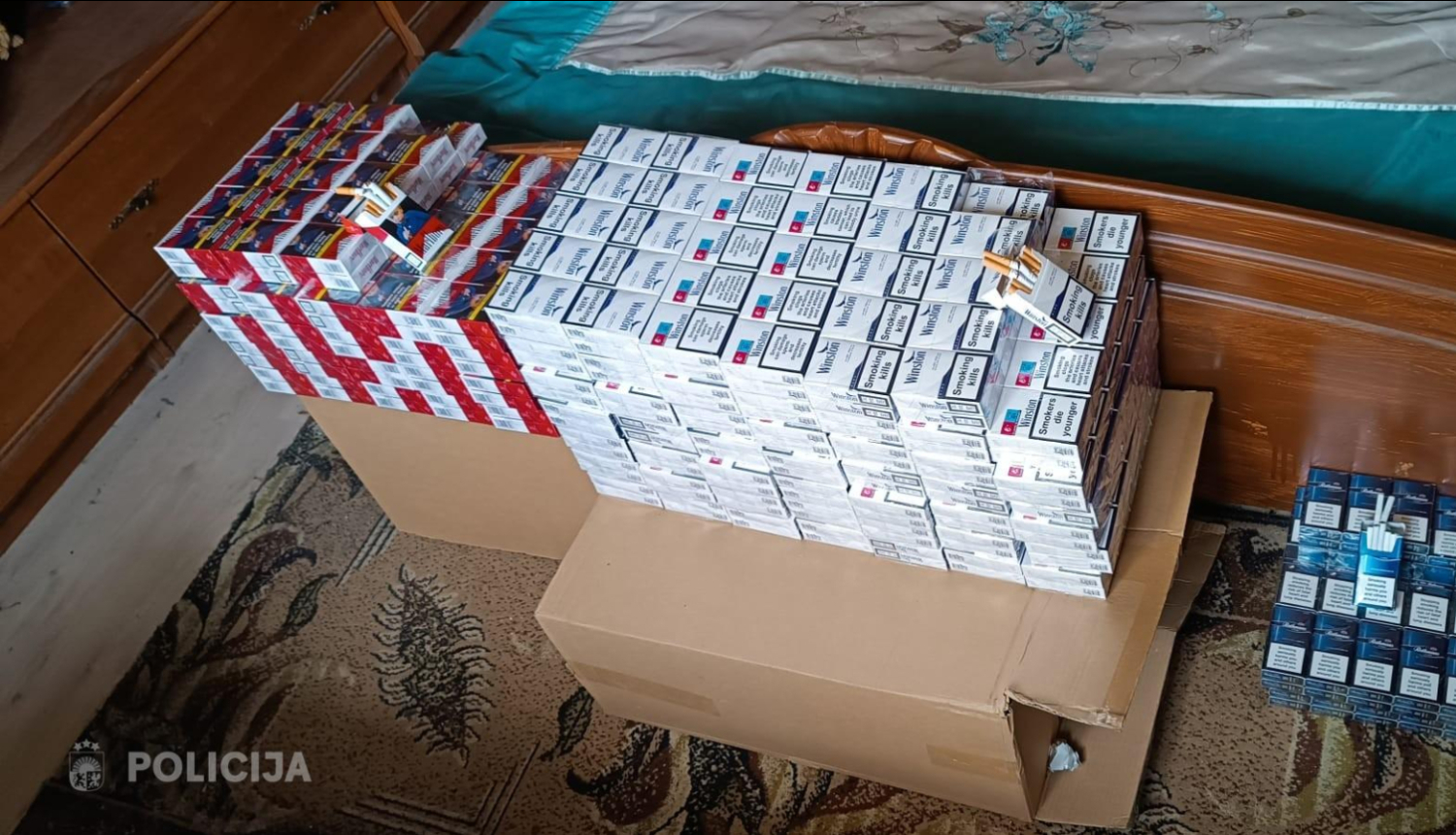 Istabā pie gultas uz kartona kastēm sakrauts liels daudzums cigarešu paciņu dažādos iepakojumos; daļa paciņu novietota arī atsevišķi uz grīdas