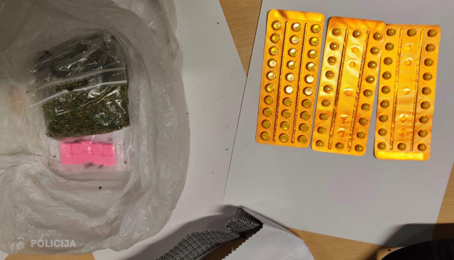 Uz galda stāv plastmasas maisiņš ar vairākiem nelieliem iepakojumiem, kuros ir zaļas augu izcelsmes vielas un rozā tabletes, blakus novietotas plāksnītes ar tabletēm