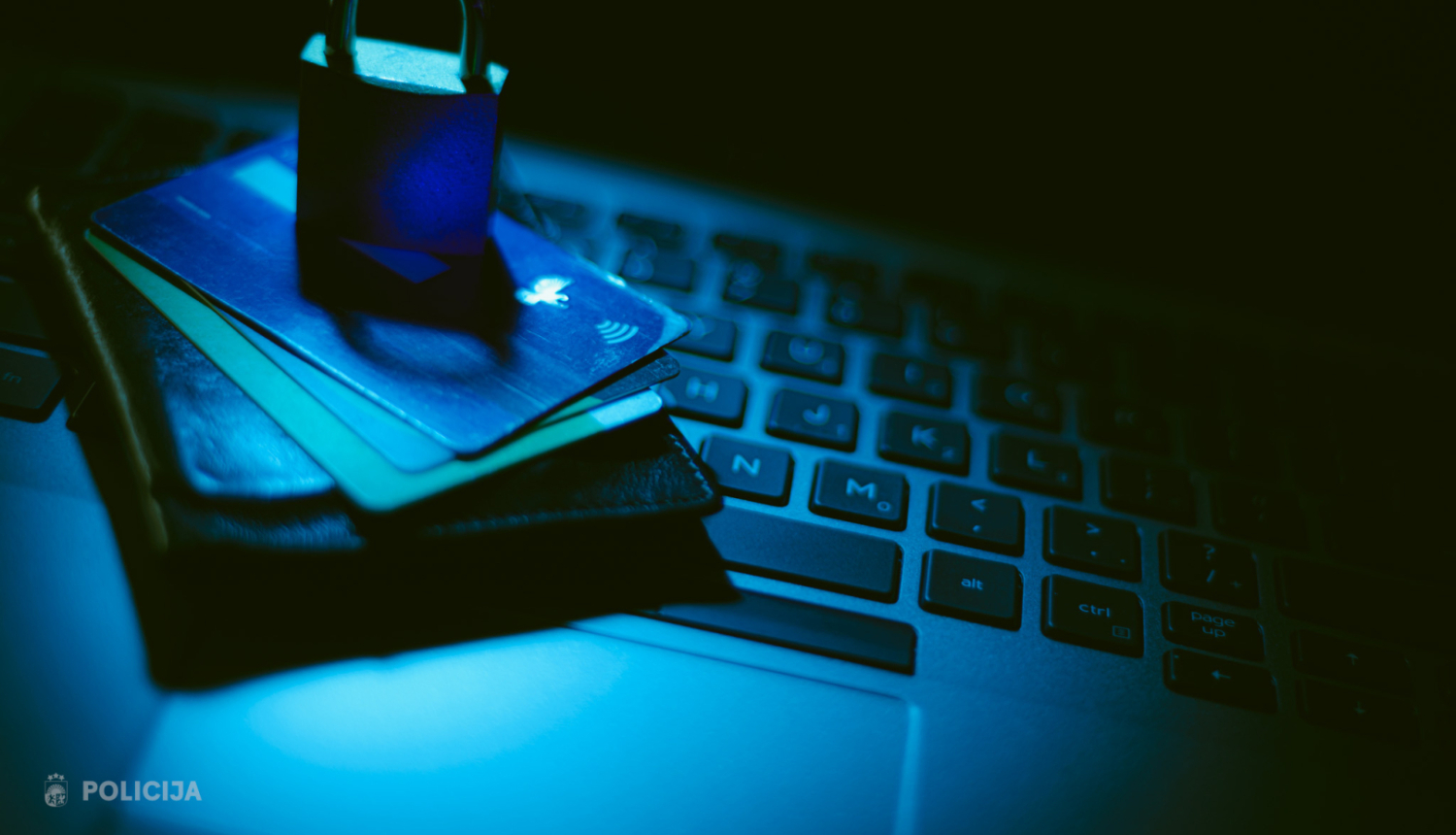 Tumšā zilganā apgaismojumā uz klēpjdatora tastatūras novietots maks ar bankas kartēm, uz kurām uzlikta piekaramā slēdzene, simboliski attēlojot finanšu un datu drošību.