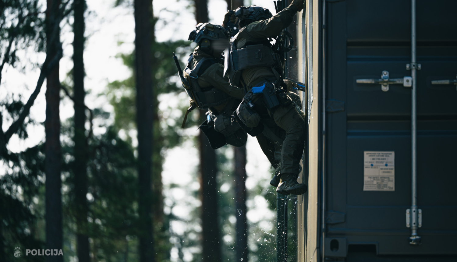 Divi Valsts policijas specvienības darbinieki taktiskajā ekipējumā treniņa laikā iekļūst ēkā mežainā apvidū