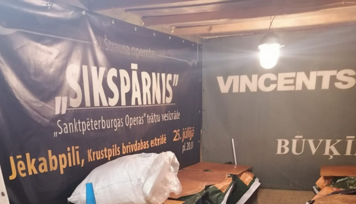 Zilas plastmasas mucas, pārsegtas ar finiera gabaliem, priekšplānā alumīnija kanna, fonā pie sienas operas izrādes "Sikspārnis" platāts un uzņēmuma "Vincents" reklāma  