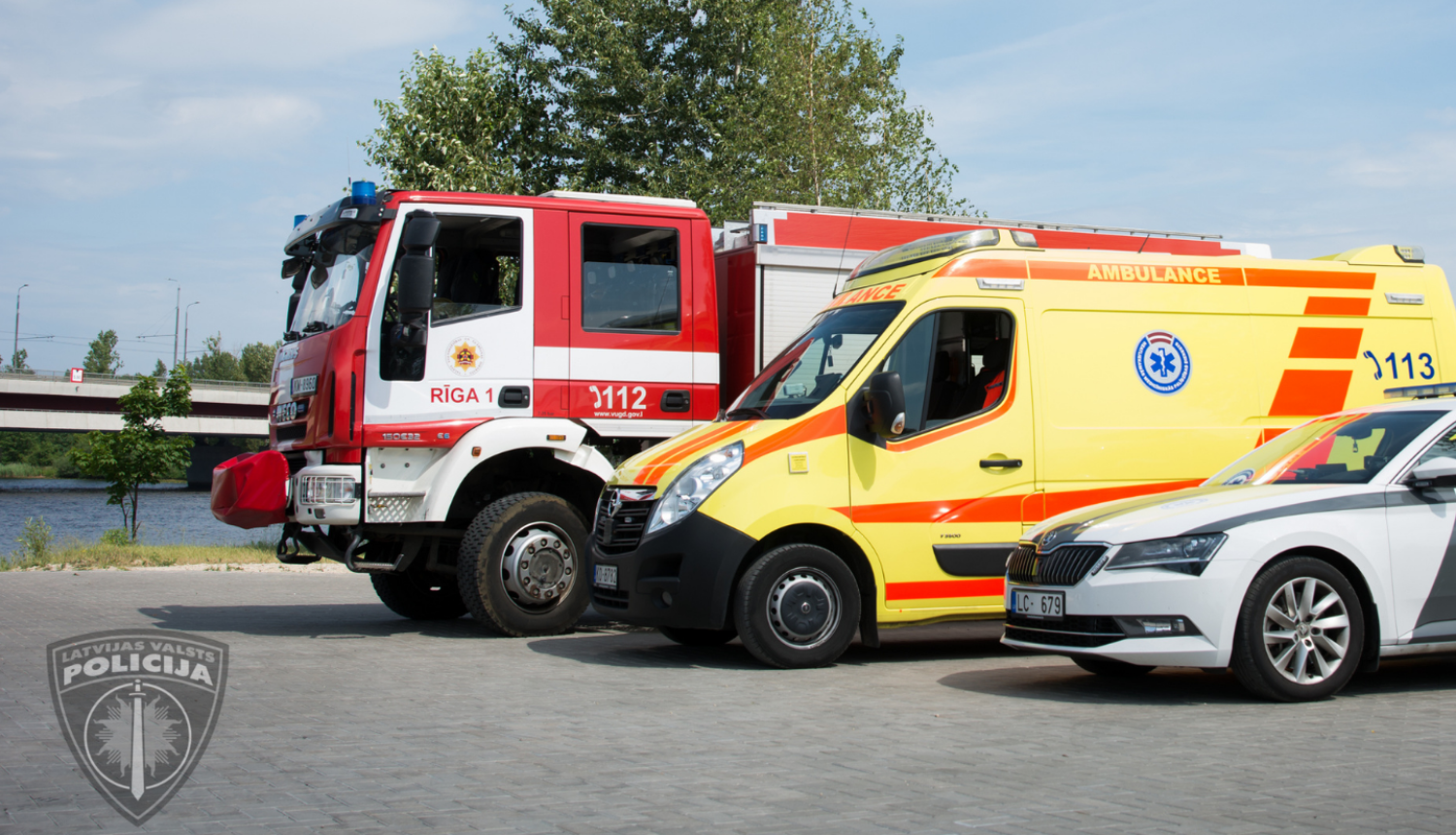 Pie upes novietoti stāvēšanai cits citam blakus operatīvie transportlīdzekļi, priekšplānā policijas mašīna, pa vidu neatliekamās medicīniskās palīdzības dienesta mašīna, tālāk valsts ugunsdzēsības un glābšanas dienesta mašīna