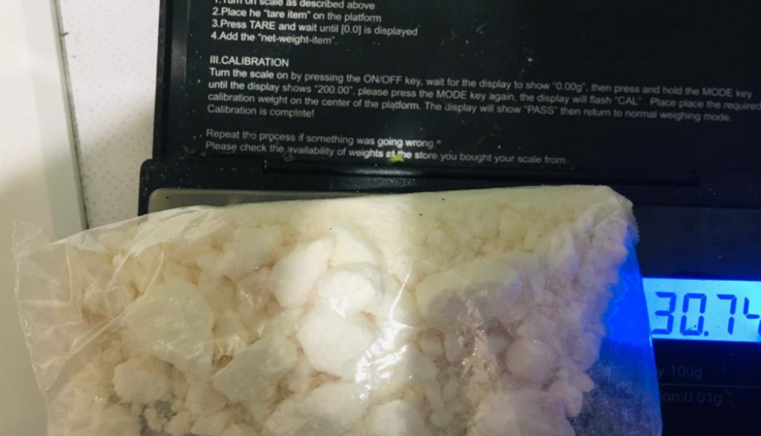Caurspīdīgā plastmasas maisiņā balta viela uzlikta uz elektroniskajiem svariem, kas rāda 30,74 gramus
