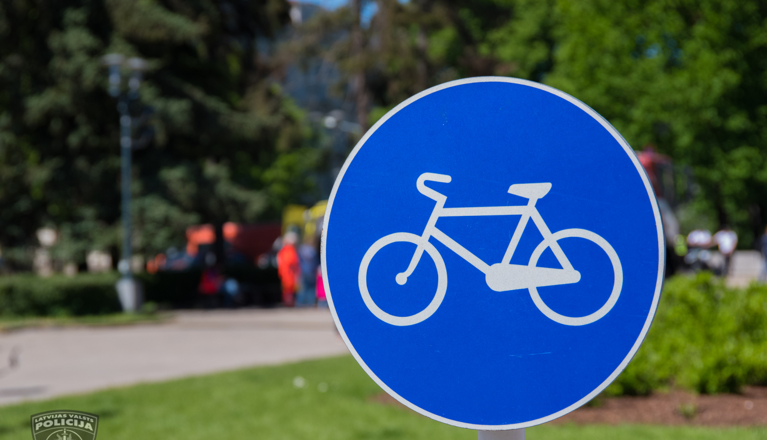 Ceļa zīme, kas norāda riteņbraucēju ceļu, fonā redzams parks