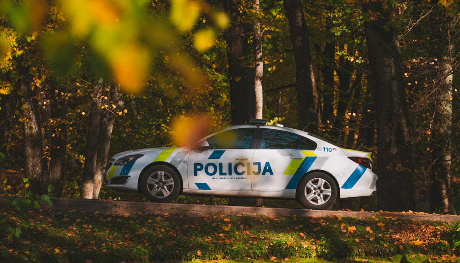 Policijas automašīna stāv uz ceļa, visapkārt koki, krāsainās rudens lapās