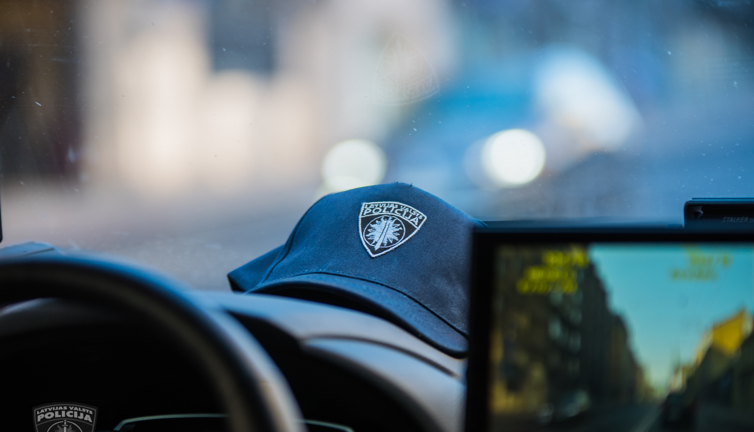 Policijas automašīnas iekšpuse, redzama stūre, daļa no ekrāna, uz priekšējā paneļa uzlikta policista cepure