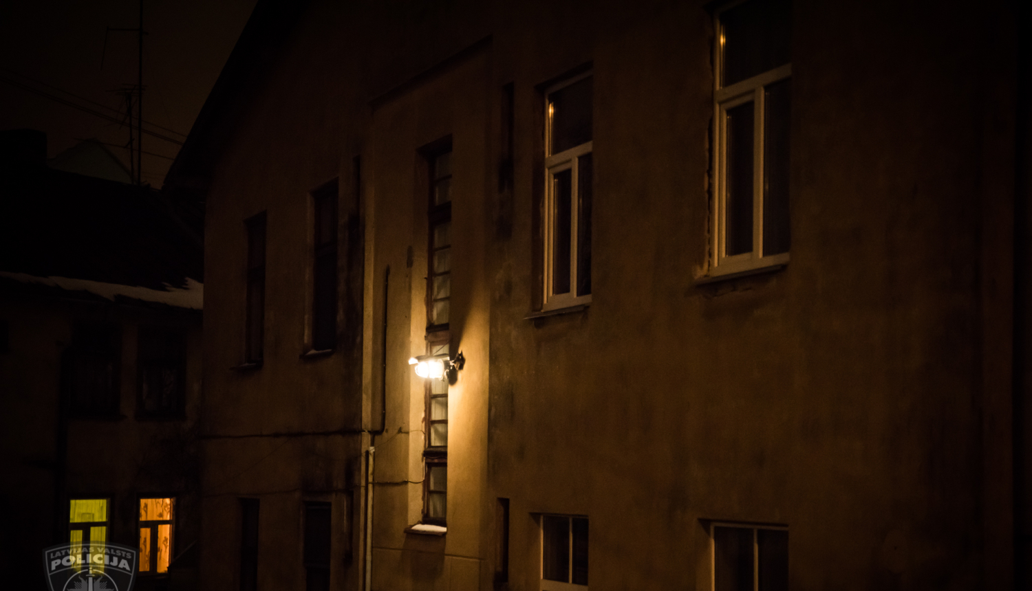 Divas divstāvu ēkas tumsā. Pie vienas virs kāpņu telpas ieejas durvīm spīd gaisma. Otrai mājai apgaismoti divi logi.