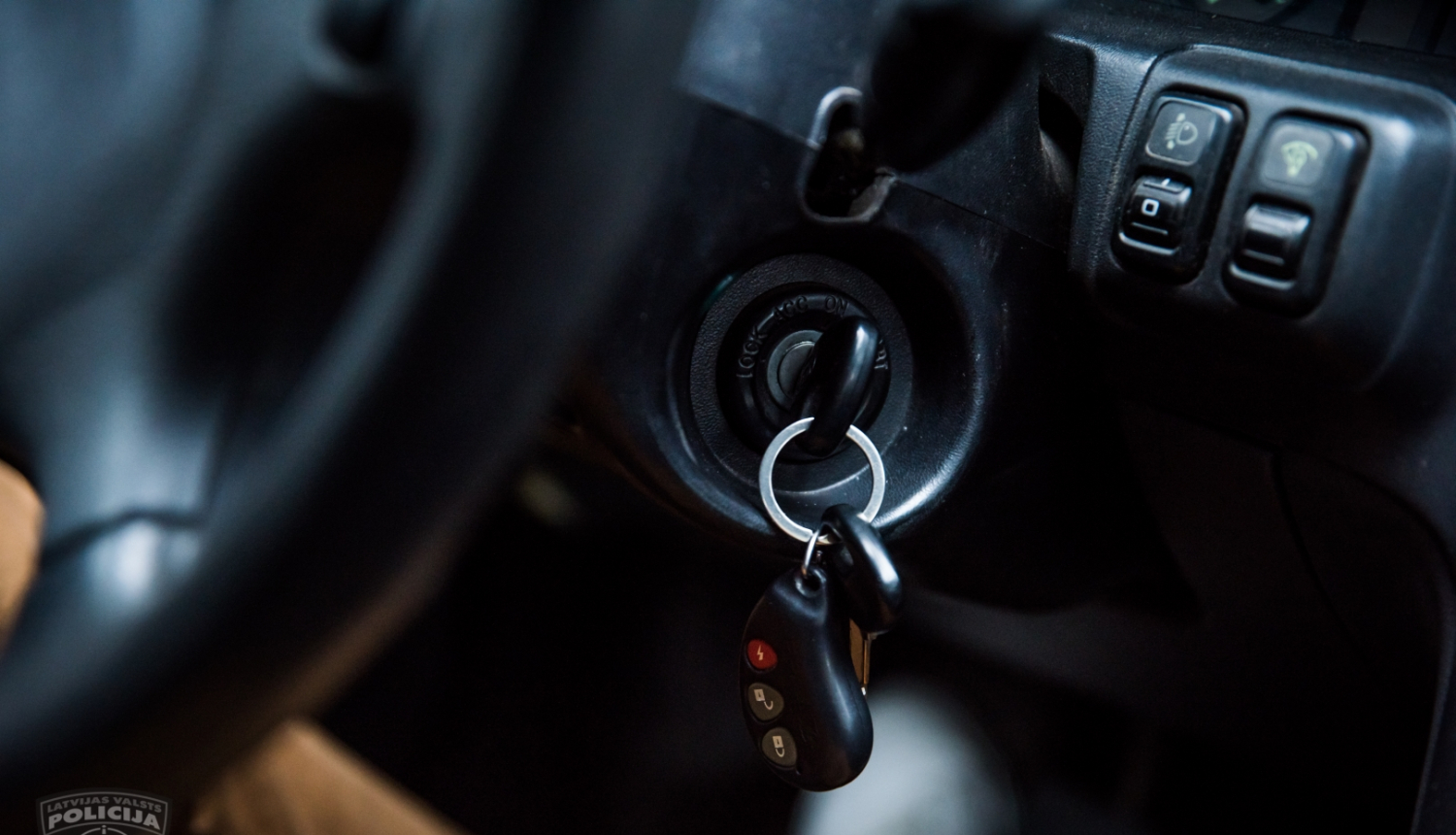 Attēlā redzama daļa no automašīnas stūres melnā krāsā, melns priekšējais panelis un aizdedzē ievietota melna atslēga