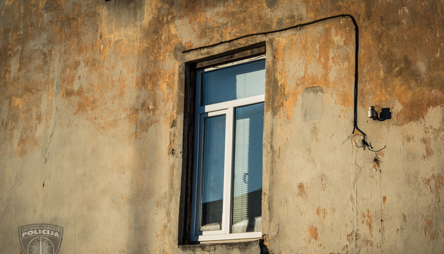 Mājas siena, viens logs ar horizontālajām žalūzijām, stūrī zem loga izdrupusi iedobe sienā