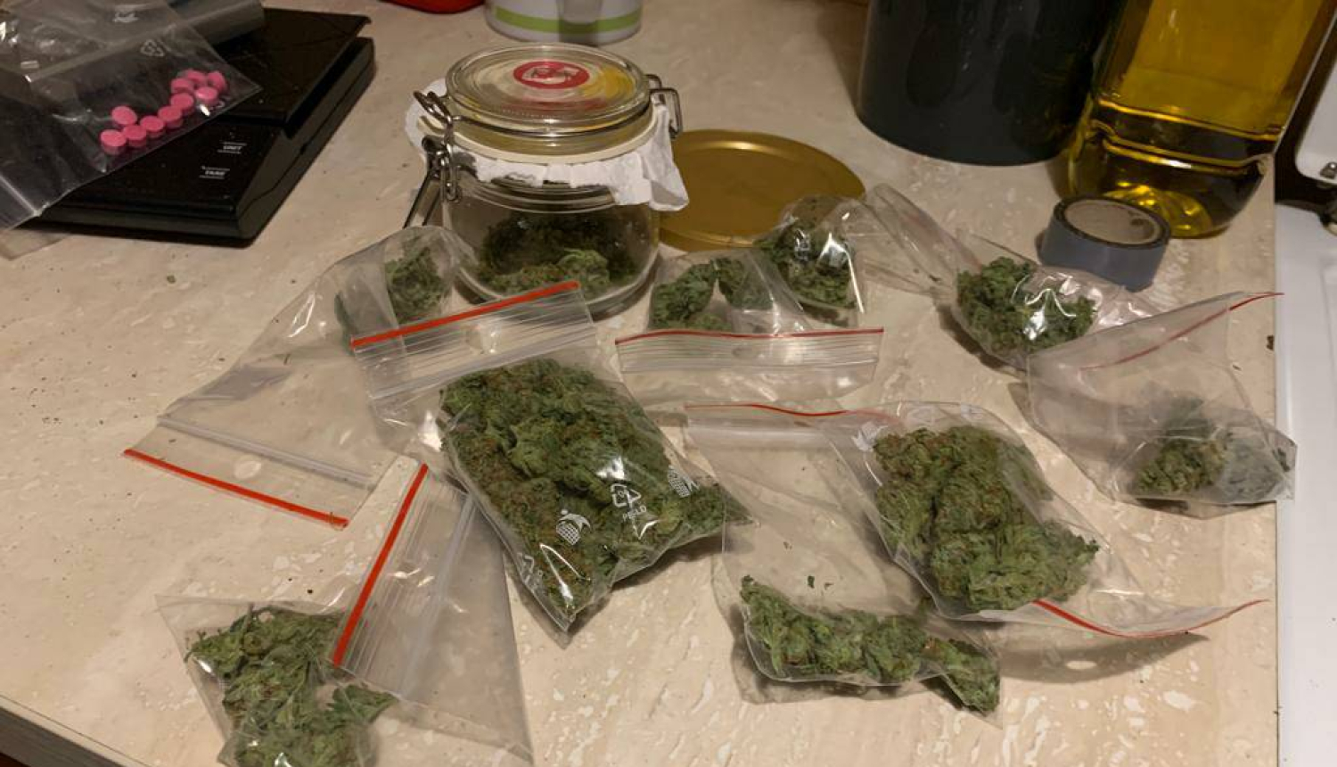 Uz galda caurspīdīgi plastikāta maisiņi ar marihuānu un uz svariem maisiņš ar rozā tabletēm