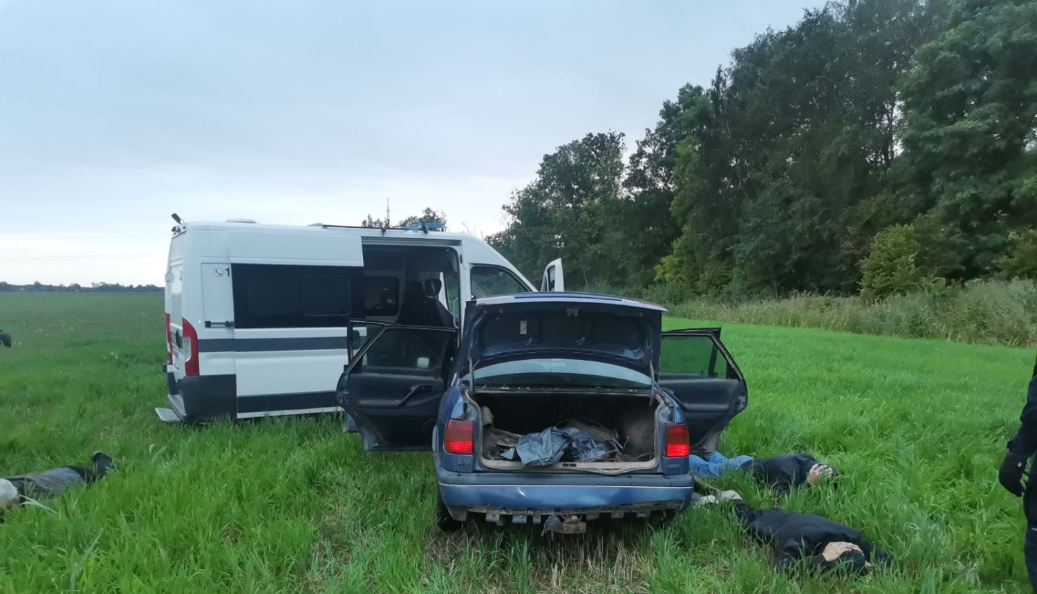 Policijas automašīna piebraukusi priekšā vieglam auto, kam visas durvis atvērtas, arī bagāžas nodalījums. Blakus automašīnai zālē guļ 3 vīrieši ar seju pret zemi