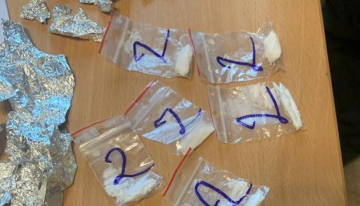 Uz galda vairāki caurspīdīgi plastmasas maisiņi, kuros iekšā balta pulverveida viela, un folijas loksnes