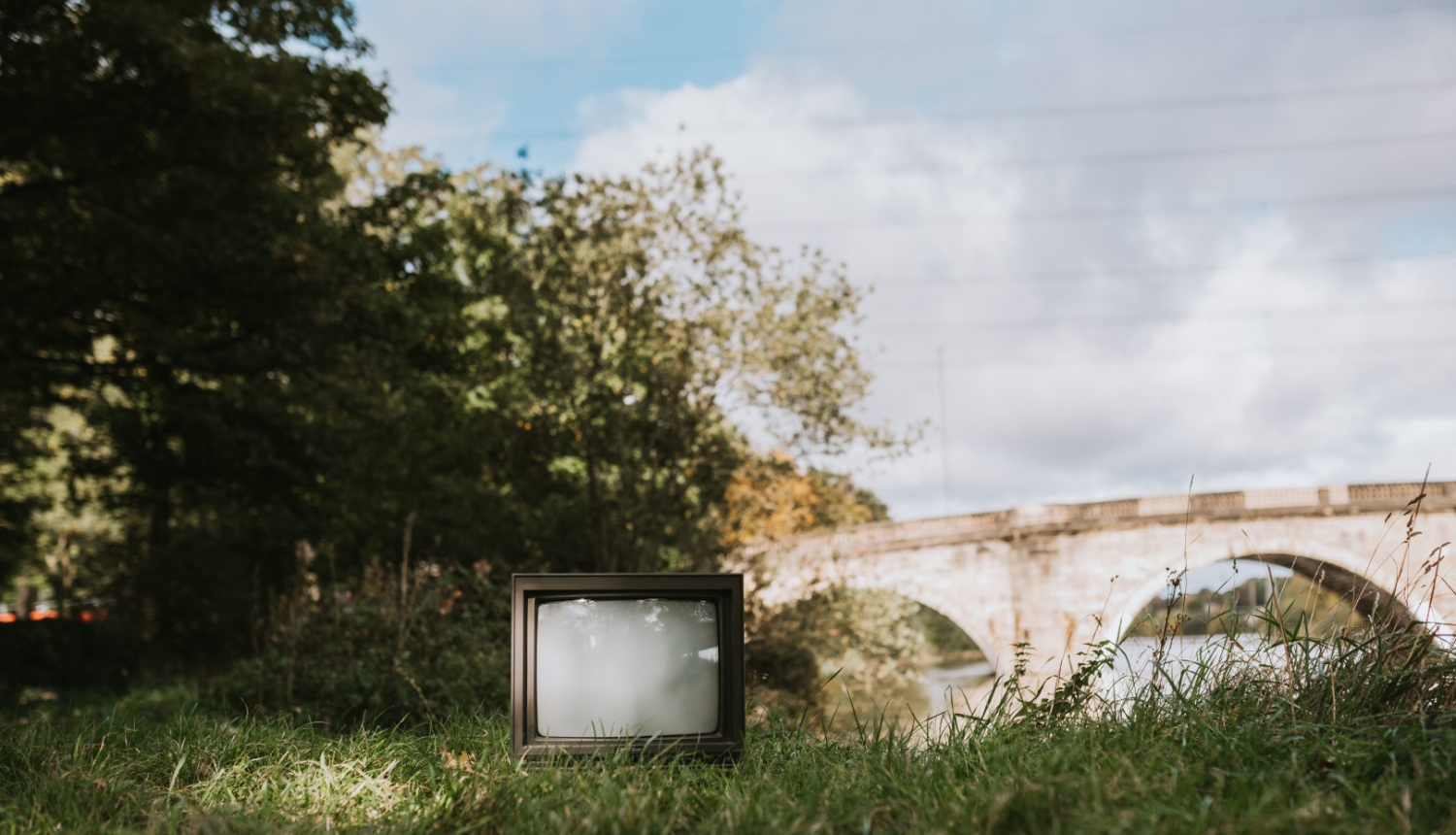 Vecs televizors novietots zālē, fonā redzami krūmi un akmens tilts ar arkām; saulaina diena; vasara