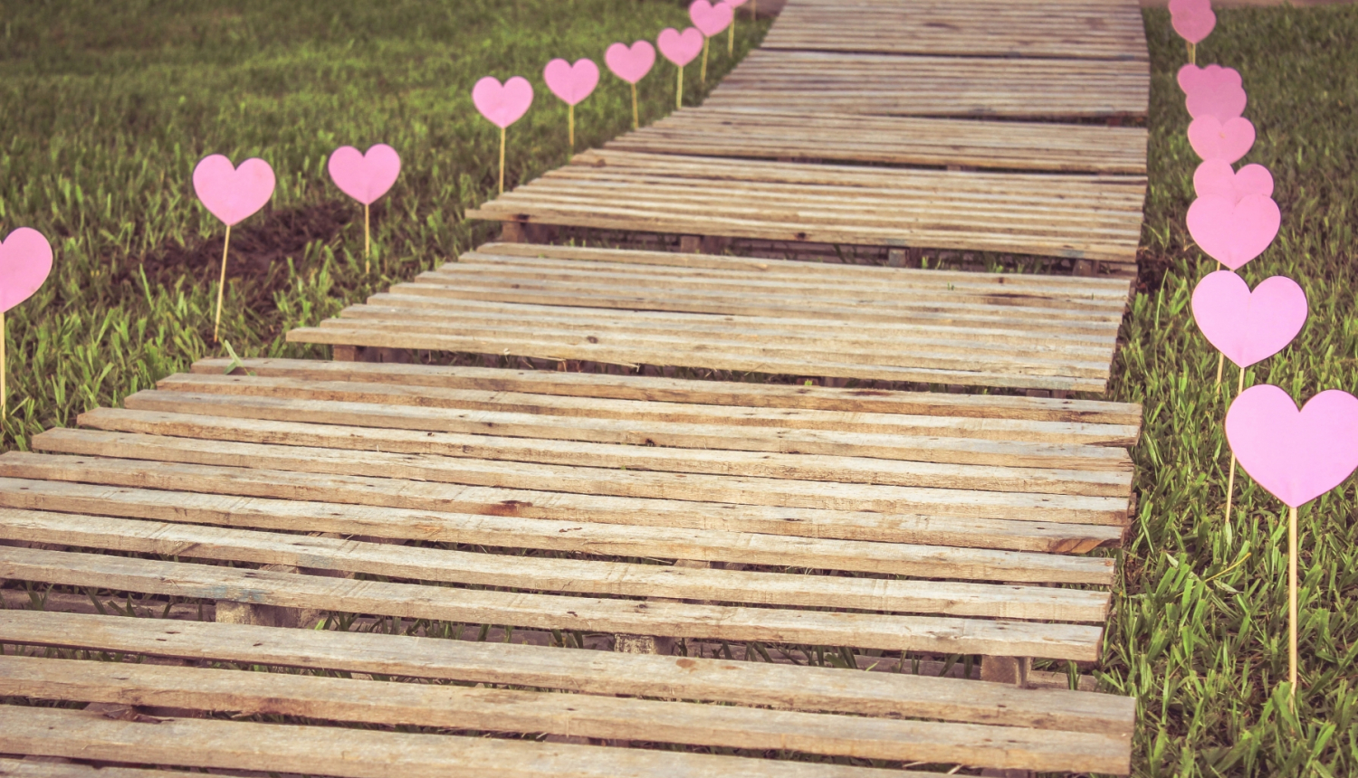 Zaļā zālē izveidots celiņš no koka dēlīšiem, kur abās malās zemē iespraustas pie irbulīša pielīmētas rozā papīra sirsniņas