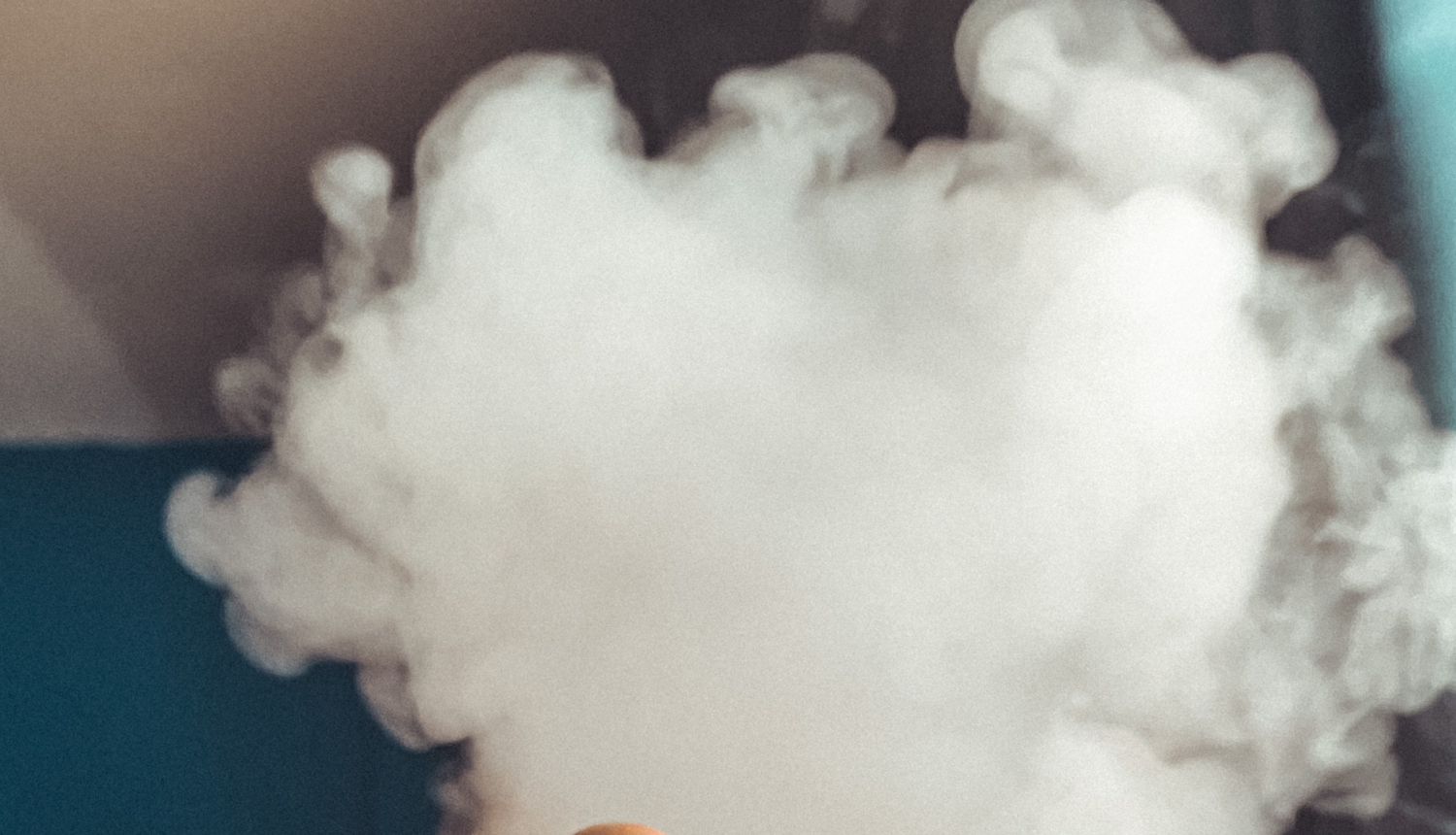 Cilvēka rokā tiek turēta melna elektroniskās smēķēšanas ierīce, seju sedz dūmu mākonis