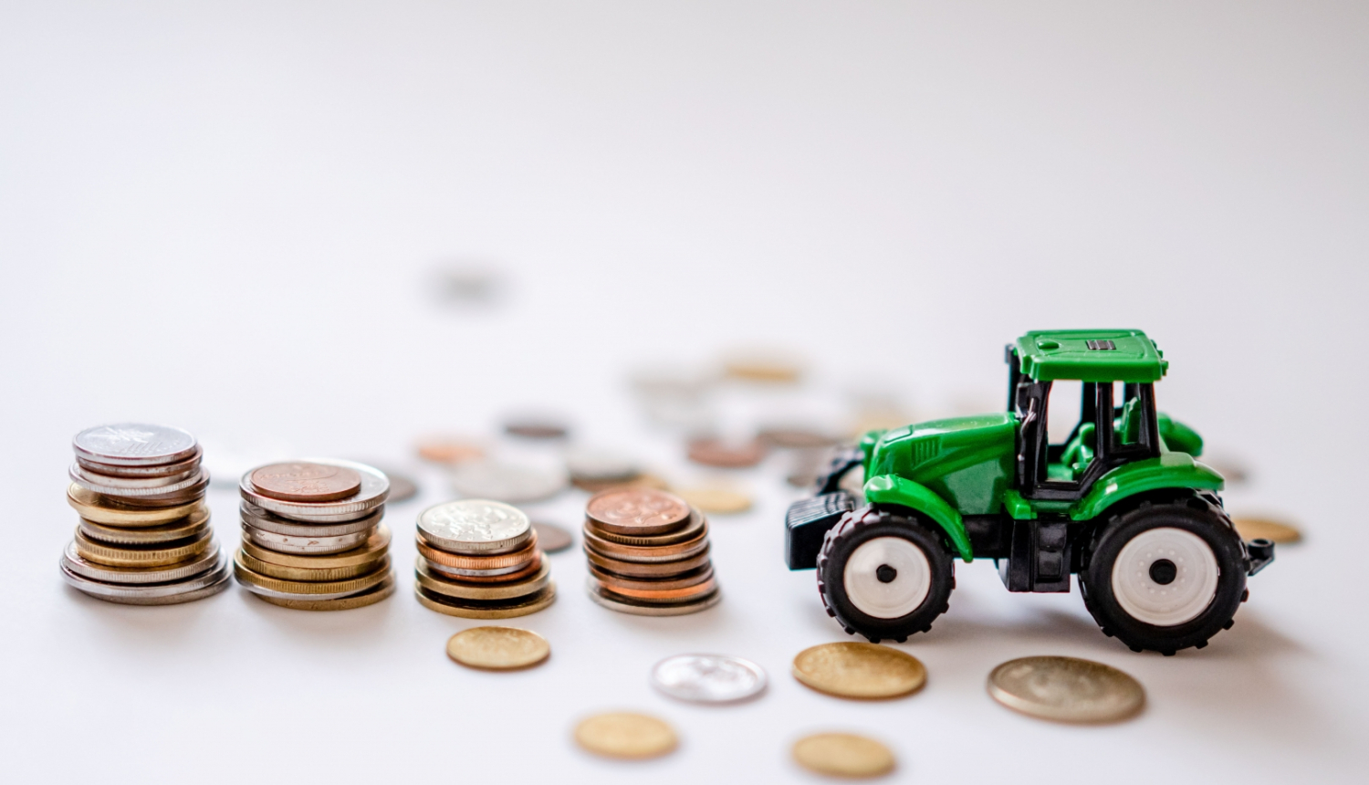Zaļš rotaļu traktors, kuram priekšā četras monētu kaudzītes un atsevišķas monētas izbārstītas pa gaišu galda virsmu