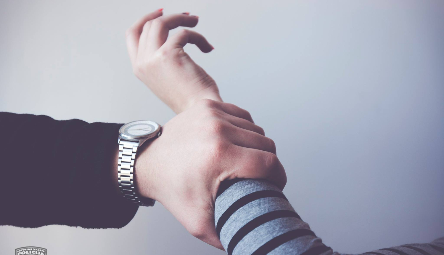 Vīrieša roka sagrābusi kādas citas personas roku. Vīrietim uz rokas pulkstenis, tumša gara piedurkne. Otras personas apģērbs - pelēki melna svītraina gara piedurkne.