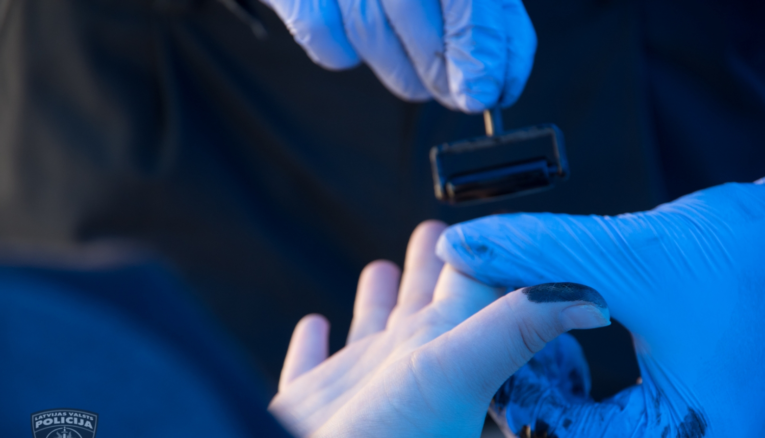 Eksperta rokas zilos gumijas cimdos un kādas personas roka, no kuras tiek noņemti pirkstu nospiedumi