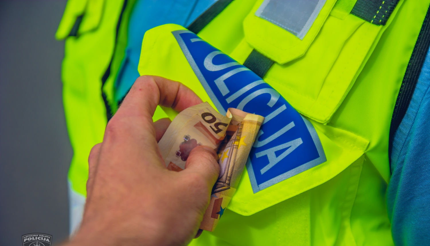 Plaukstā redzami 50 eiro, ko cenšas ielikt policista formas apģērba vestes priekšējā kabatā