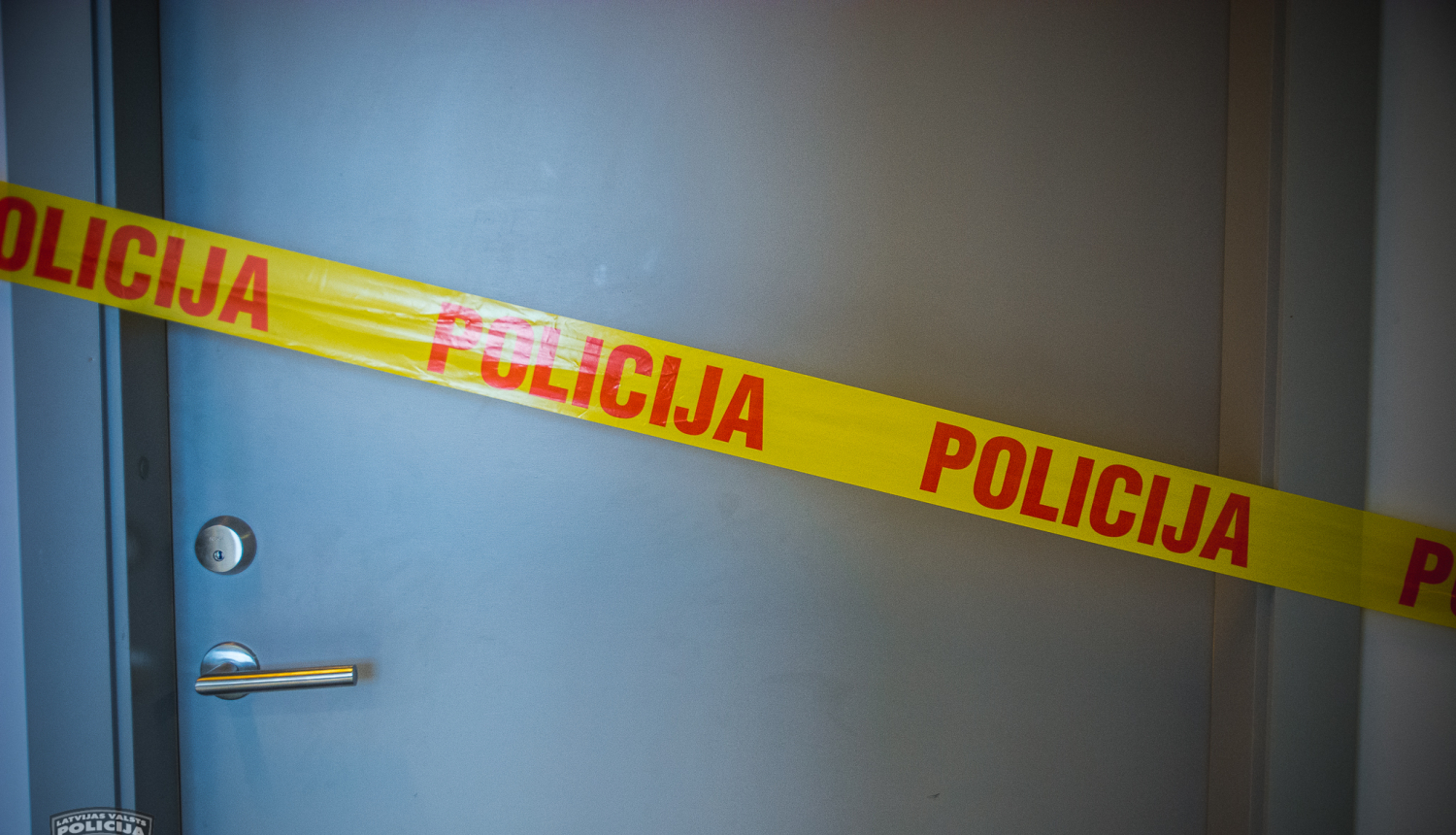 Pelēkas durvis, kurām priekšā novilkta dzeltena lenta, uz kuras ar sarkaniem burtiem uzrakstīts "Policija"