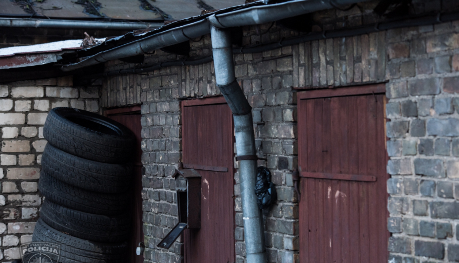 Ķieģeļu ēka, trīs brūnas koka ārdurvis, vienām durvīm priekšā sakrautas automašīnu riepas